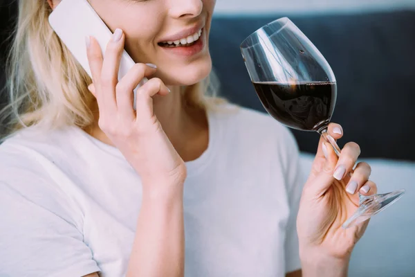 Vista recortada de la mujer sonriente sosteniendo un vaso de vino y hablando en el teléfono inteligente en casa - foto de stock