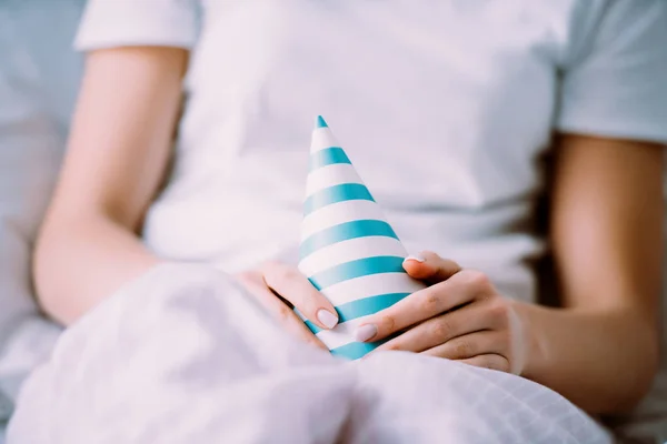 Cropped view of woman in pajamas holding party hat while celebrating birthday in bed alone — Stock Photo