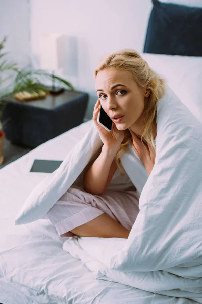 Mujer triste cubierta de manta mirando a la cámara y hablando en el teléfono inteligente en la cama en casa - foto de stock