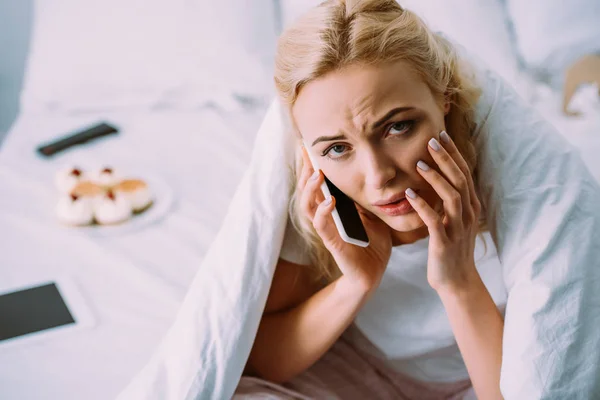 Sad woman covered in blanket looking at camera and talking on smartphone in bed at home — Stock Photo