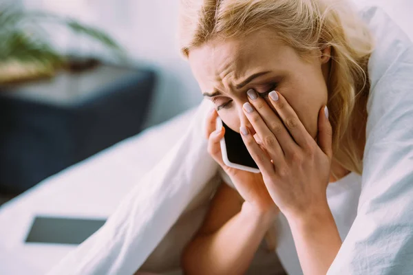 Traurige Frau in Decke gehüllt, telefoniert auf Smartphone und weint zu Hause im Bett — Stockfoto