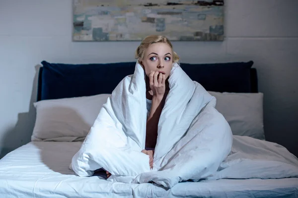 Scared woman covered in blanket biting nails in bed at home and looking away — Stock Photo