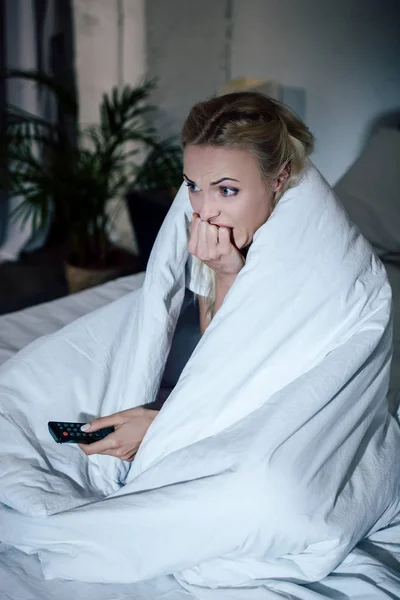 Scared woman covered in blanket holding remote control, biting nails and watching tv in bed at home — Stock Photo