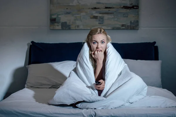 Scared woman covered in blanket holding remote control, biting nails and watching tv in bed at home — Stock Photo