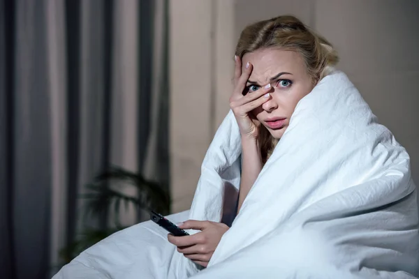Verängstigte Frau deckt Gesicht mit der Hand ab, während sie zu Hause im Bett fernsieht — Stockfoto