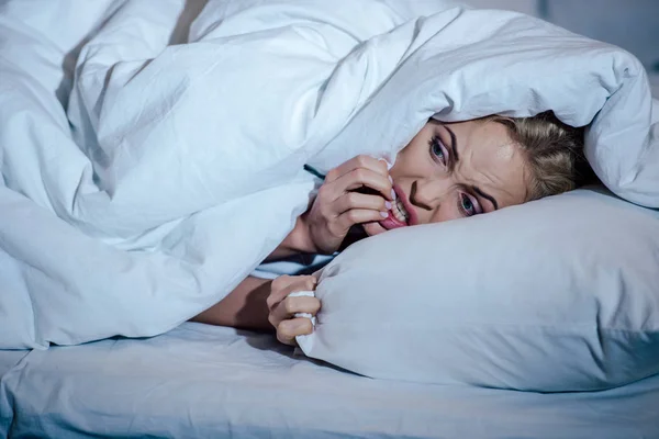 Mujer cubierta de manta mordiendo clavos en la cama en casa - foto de stock