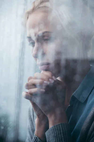 Mulher adulta triste com as mãos dobradas em casa através da janela com gotas de chuva — Fotografia de Stock