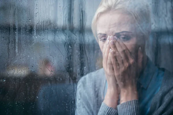 Triste donna adulta piangendo e coprendo il viso con le mani a casa attraverso la finestra con gocce di pioggia — Foto stock