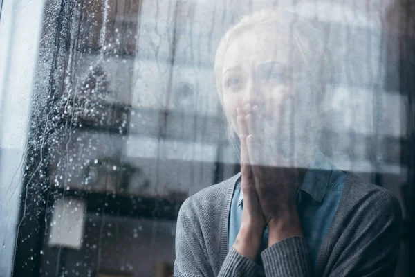 Sad adult woman with praying gesture crying at home through window with raindrops — Stock Photo