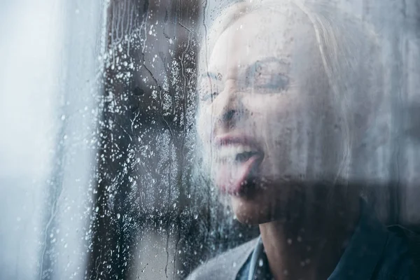 Foco seletivo de mulher adulta irritada colando a língua para fora e gritando em casa através da janela com gotas de chuva — Fotografia de Stock