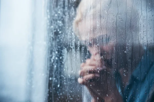 Traurige erwachsene Frau mit gefalteten Händen zu Hause durch Fenster mit Regentropfen — Stockfoto