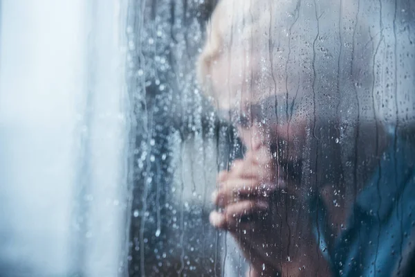 Sad adult woman with folded hands at home through window with raindrops — Stock Photo