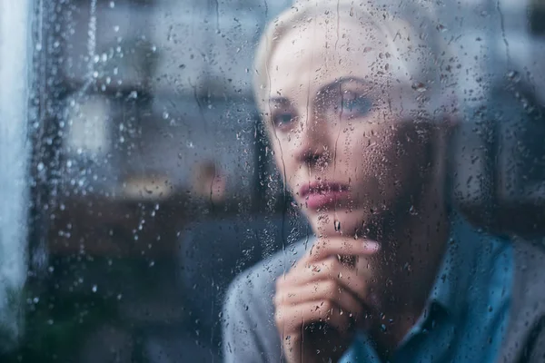 Грустная задумчивая взрослая женщина, прикасающаяся лицом к дому через окно каплями дождя — стоковое фото