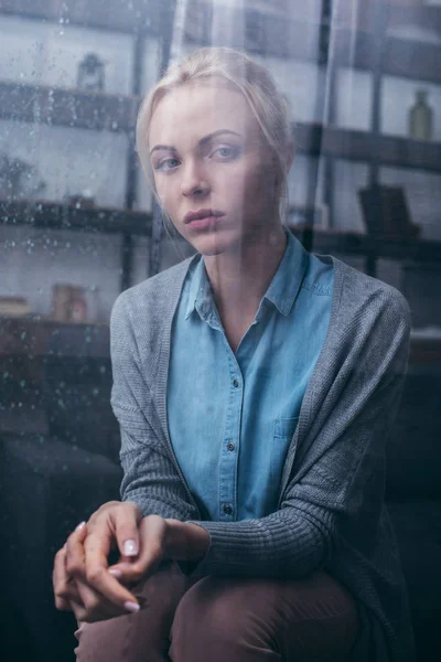 Triste donna adulta seduta con le mani giunte e guardando la fotocamera a casa attraverso la finestra con gocce di pioggia — Foto stock