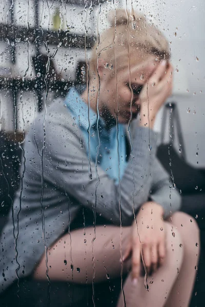Femme adulte triste touchant la tête et ayant mal à la tête à la maison par la fenêtre avec des gouttes de pluie — Photo de stock