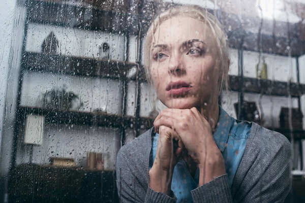 Mulher adulta triste com as mãos dobradas olhando para casa através da janela com gotas de chuva — Fotografia de Stock