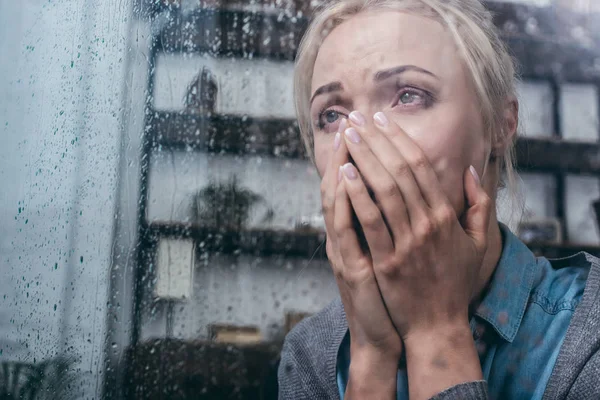 Triste mujer adulta llorando y cubriendo la cara con las manos en casa a través de la ventana con gotas de lluvia - foto de stock