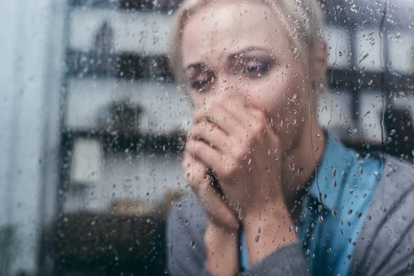 Donna adulta depressa con le mani piegate a casa attraverso la finestra con gocce di pioggia — Foto stock