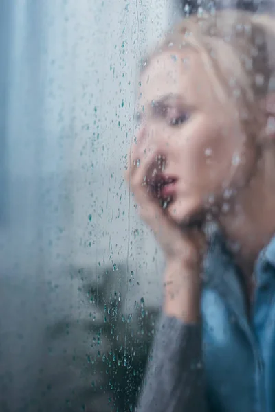 Selektiver Fokus der traurigen Frau, die Gesicht mit der Hand durch Fenster mit Regentropfen und Kopierraum bedeckt — Stockfoto