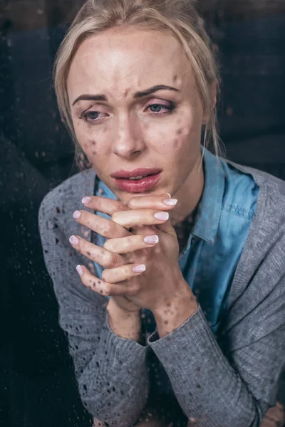 Chateado mulher adulta com as mãos dobradas chorando em casa através da janela com gotas de chuva — Fotografia de Stock