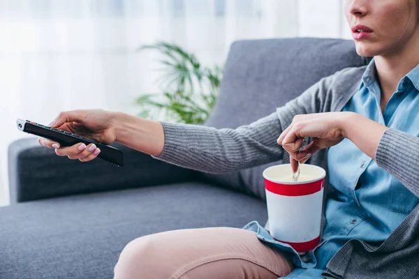 Cropped view of woman sitting on couch with ice cream and remote control while watching tv — Stock Photo
