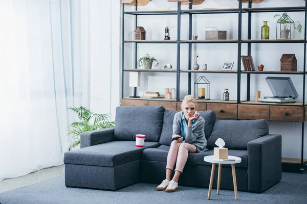 Sad woman sittng on couch with bucket of ice cream and tissue box while watching tv in living room — Stock Photo