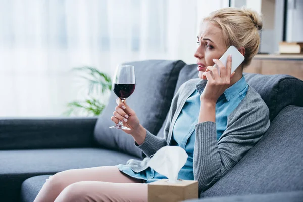 Засмучена жінка сидить на дивані, тримає келих червоного вина і розмовляє на смартфоні вдома — стокове фото