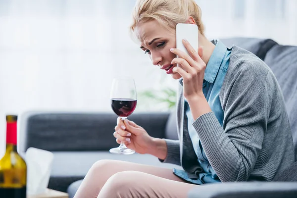 Chateado mulher sentado no sofá, segurando copo de vinho tinto e falando no smartphone em casa — Fotografia de Stock