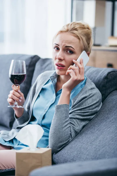 Femme assise sur le canapé, regardant la caméra, tenant un verre de vin rouge et parlant sur smartphone à la maison — Photo de stock