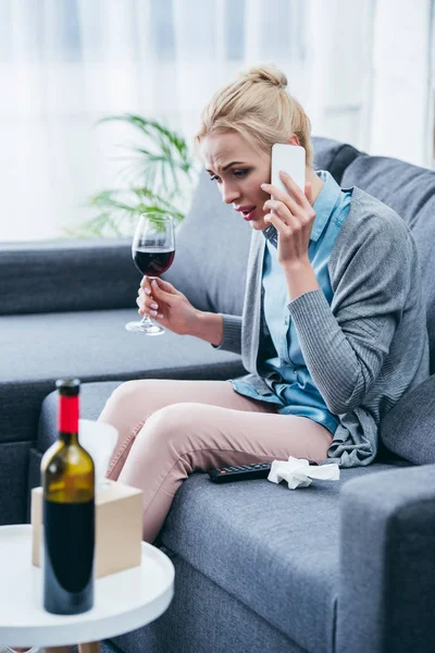 Woman sitting on couch, holding glass of red wine and talking on smartphone at home — Stock Photo