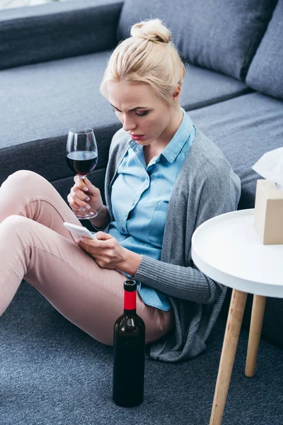 Traurige Frau hält Glas Wein in der Hand und benutzt Smartphone zu Hause — Stockfoto