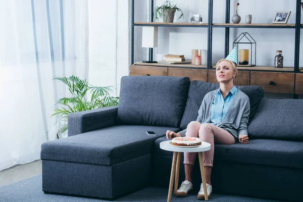 Frustrierte Frau mit Partyhut sitzt auf Couch und feiert Geburtstag allein zu Hause — Stockfoto