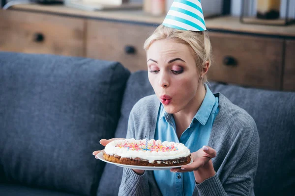 Woman in party hat blowing out candle on cake while celebrating birthday at home alone — Stock Photo