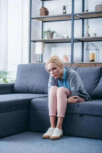 Mujer triste en ropa casual sentada en el sofá en la sala de estar - foto de stock