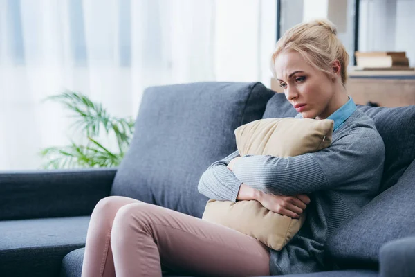Подавленная женщина сидит на диване и держит подушку дома — стоковое фото