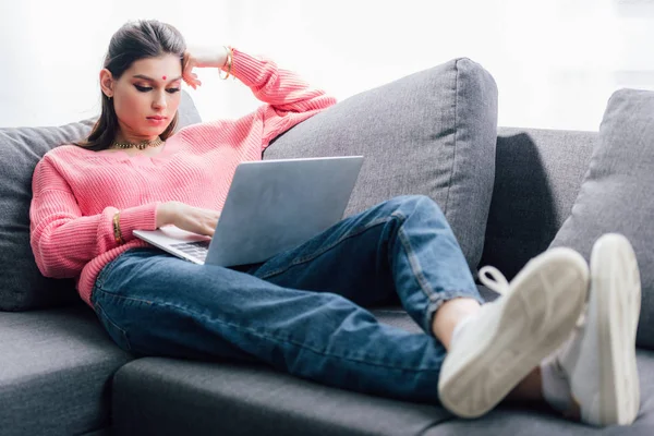 Bella donna indiana con bindi utilizzando il computer portatile mentre si trova sul divano — Foto stock