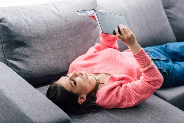 Attractive indian student with bindi reading book and lying on sofa — Stock Photo