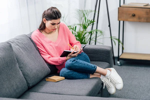 Indische Studentin schreibt in Notizbuch, während sie mit Buch auf Sofa sitzt — Stockfoto