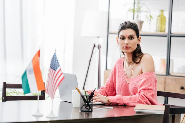 Attractive indian girl with bindi studying with laptop at table with american and indian flags — Stock Photo