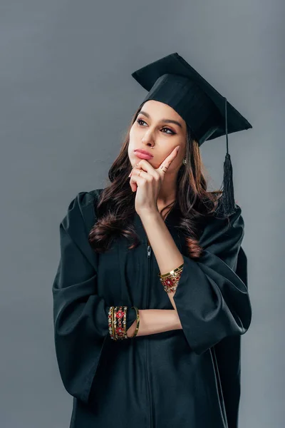 Estudiante indio pensativo en vestido académico y gorra de graduación, aislado en gris - foto de stock