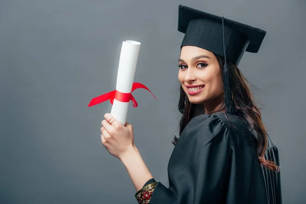 Sorridente estudante indiana em vestido acadêmico e diploma cap graduação segurando, isolado em cinza — Fotografia de Stock