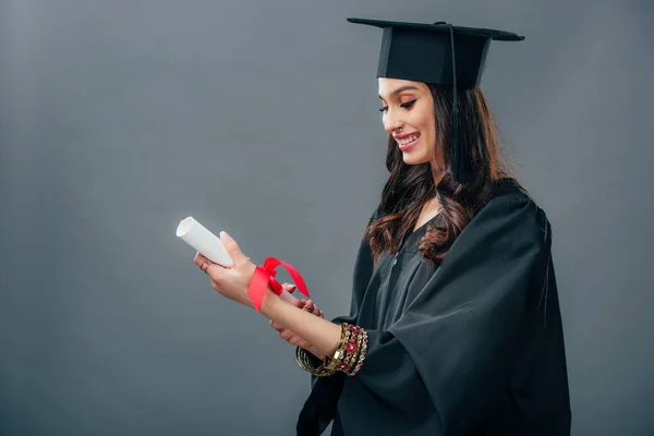 Estudante indiano feliz em vestido acadêmico e diploma chapéu de graduação segurando, isolado em cinza — Fotografia de Stock