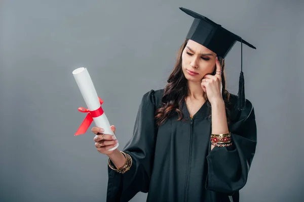 Thoughtful indian student in academic gown and graduation hat looking at diploma, isolated on grey — Stock Photo