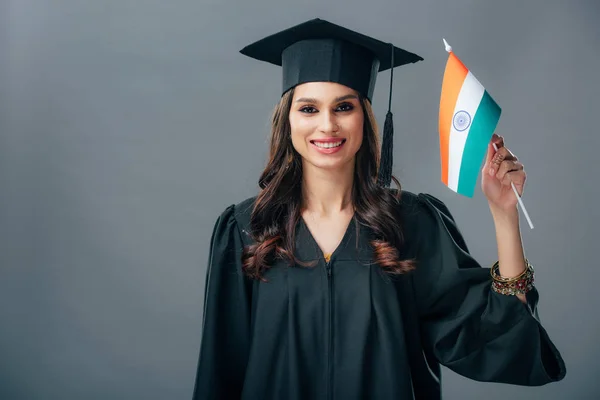 Étudiant indien souriant en robe académique et casquette de graduation tenant drapeau indien, isolé sur gris — Photo de stock