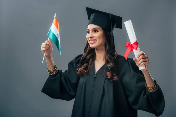 Жінка-індійська студентка в академічній сукні та випускній кепці, що тримає диплом та індійський прапор, ізольована на сірому — стокове фото