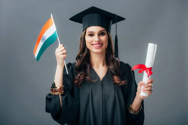 Щасливий студент в академічній сукні та випускній кепці, що тримає диплом та індійський прапор, ізольований на сірому — стокове фото
