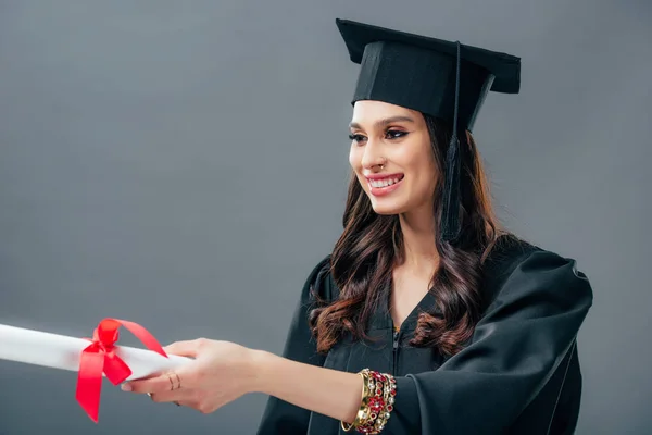 Chica sonriente en vestido académico y el sombrero de graduación haking diploma, aislado en gris - foto de stock