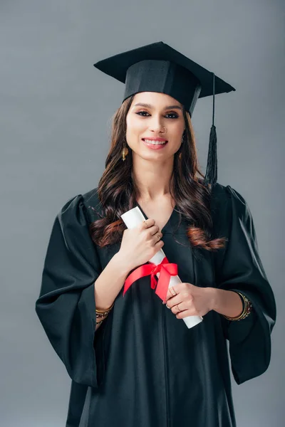 Cheerful indian woman in academic gown and graduation hat holding diploma, isolated on grey — Stock Photo