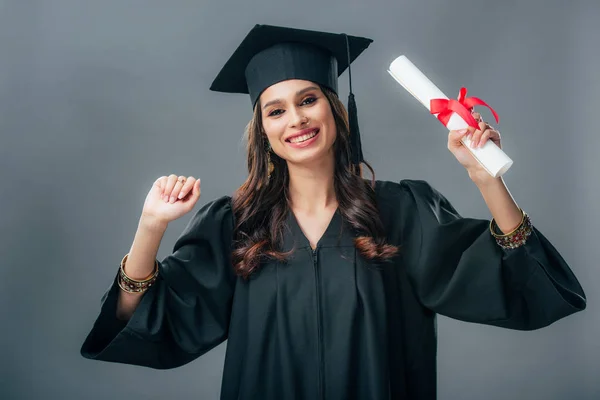 Щасливий індійський студент в академічній сукні та випускному капелюсі, який тримає диплом, ізольований на сірому — стокове фото