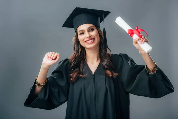 Happy indian student in academic gown and graduation hat holding diploma, isolated on grey — Stock Photo